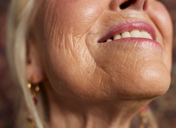 Nærbilde av den nedre delen av en persons ansikt og hals mot en uskarp bakgrunn. Personen smiler med delvis synlige tenner. Hudens naturlige struktur rundt munn, hake og hals er tydelig. Personen har små øredobber.