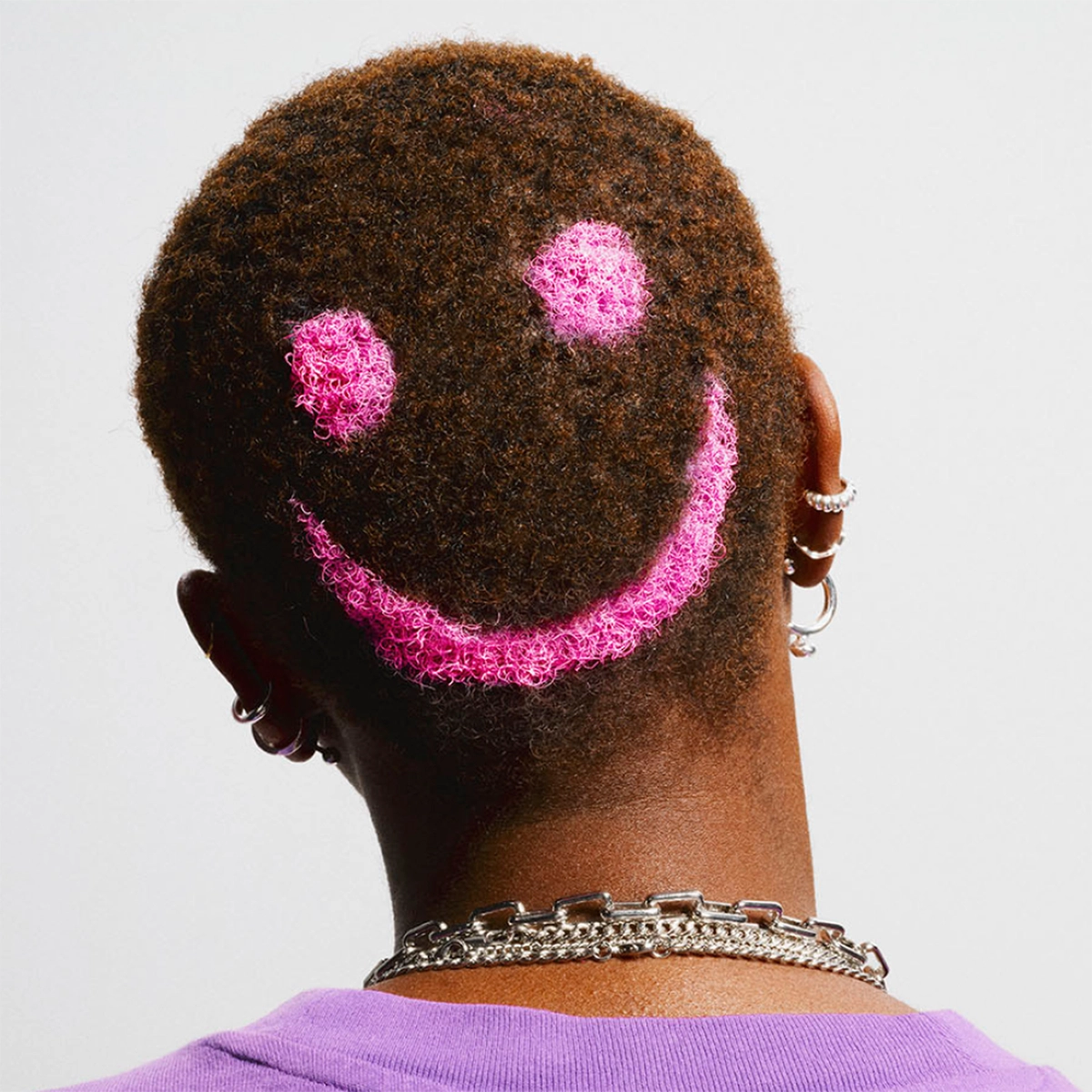 Bakhuvud på en person med mycket kort hår där ett rosa smiley-ansikte är färgat i håret, personen bär örhängen och halsband mot ljus bakgrund.