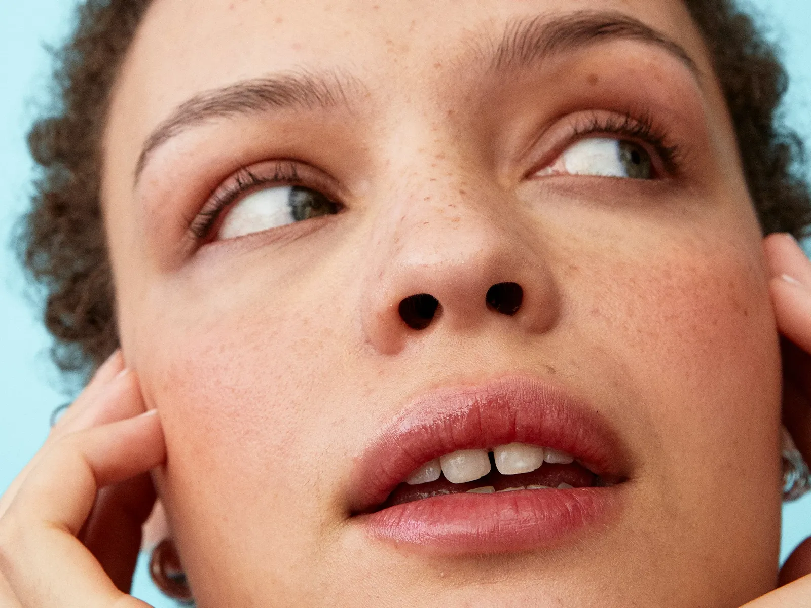 En person med krøllete hår holder hendene ved ansiktet mot en lys blå bakgrunn.