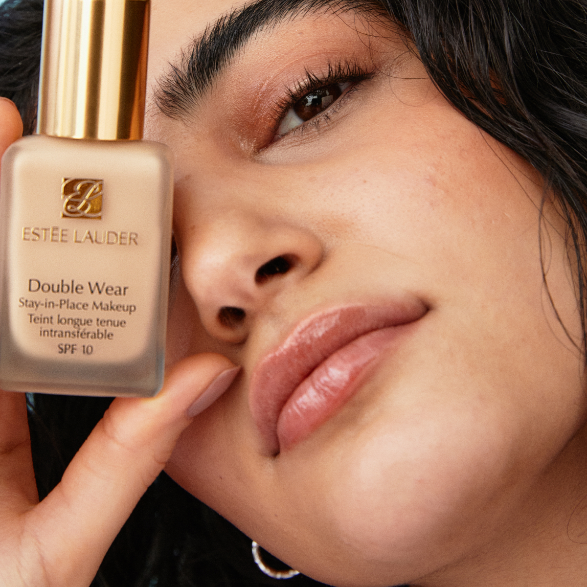 En hånd holder en flaske Estée Lauder Double Wear Stay-in-Place Makeup SPF 10. Flasken har en gullkork og teksten på flasken er tydelig synlig.