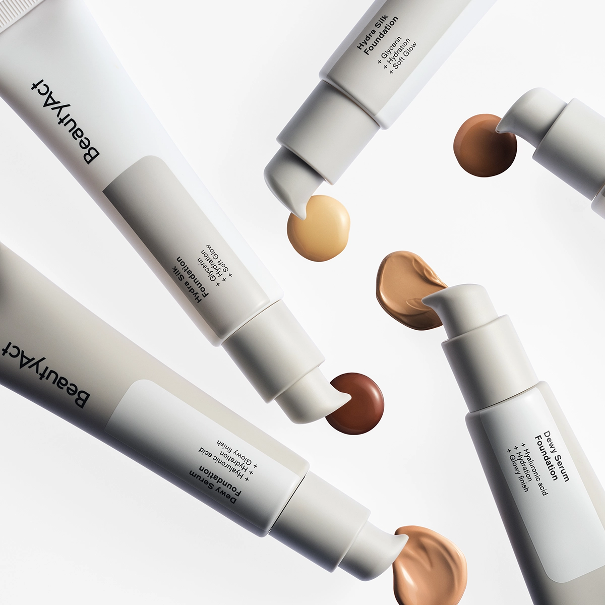 Bilde som viser forskjellige foundationprodukter fra BeautyAct. På bildet vises fem tuber med forskjellige nyanser av foundation, inkludert lys, medium og mørk. Hver tube har en pumpe som slipper ut en liten mengde foundation. Produktene er arrangert i en sirkel på en hvit bakgrunn.