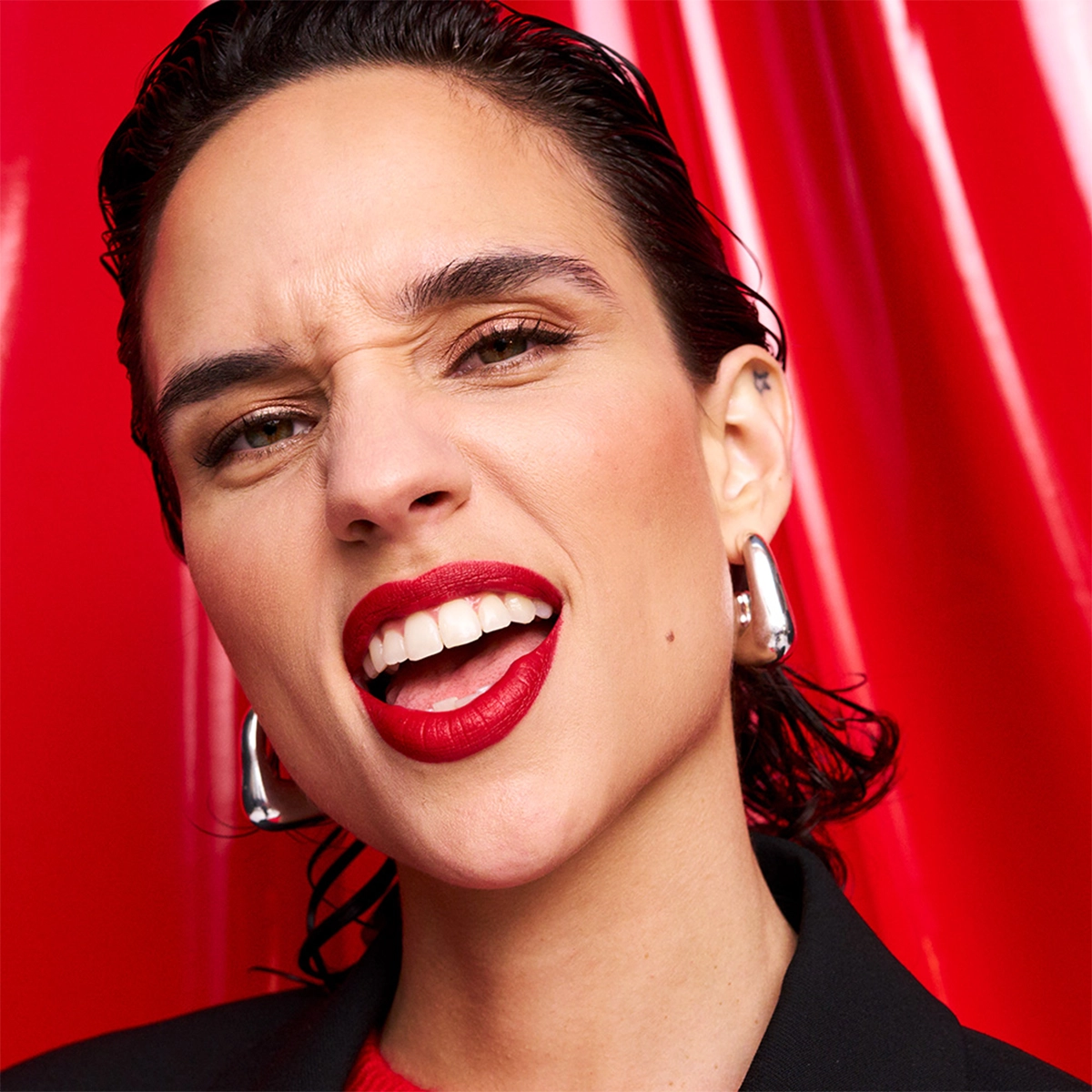 Närbild på en person med rött läppstift, bakåtkammat hår och stora silverörhängen mot en glansig röd bakgrund. En kraftfull och självsäker look som förmedlar festlig glamour och energi.