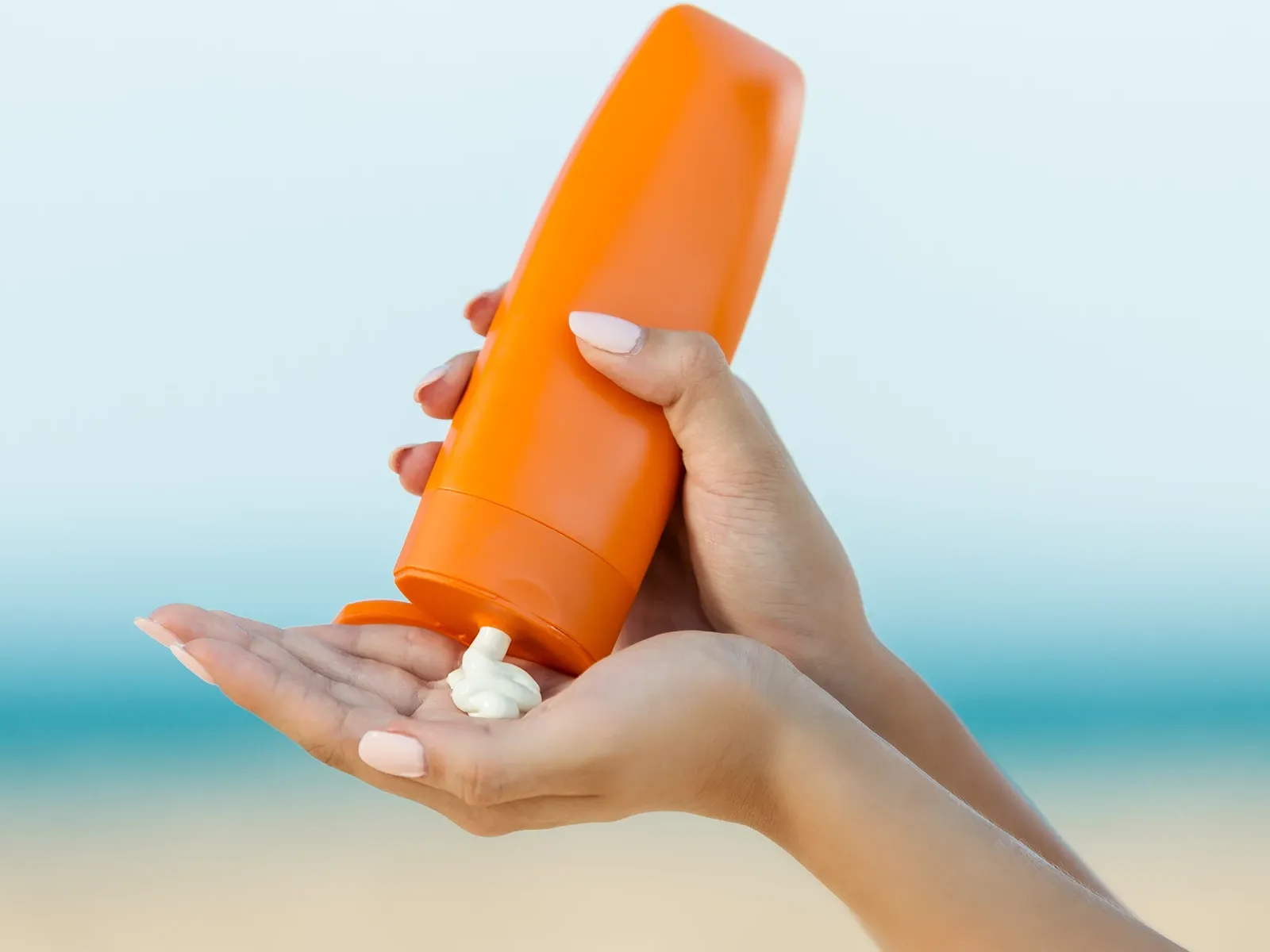 En person håller en orange flaska och applicerar solkräm på sin hand vid stranden.