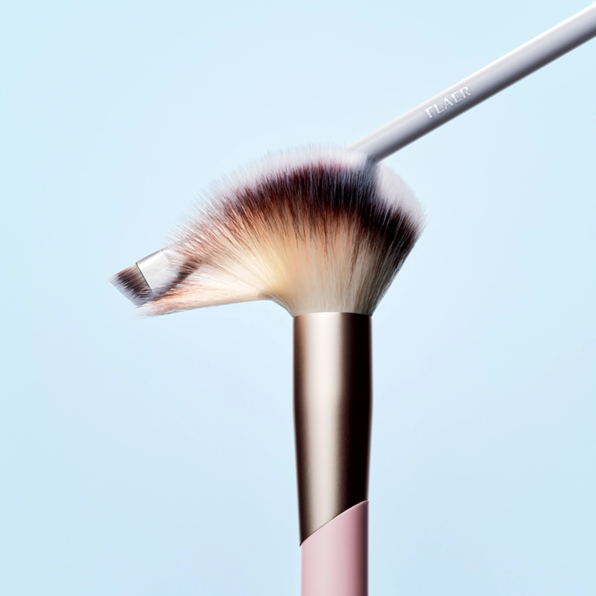 Kaksi meikkisivellintä vaaleansinistä taustaa vasten, suuri pöyheä sivellin beigeillä ja tummilla harjaksilla sekä metallinhohtoinen kuparinvärinen varsi, ja pienempi viistopäinen sivellin asetettuna sen päälle. Lähikuva, jossa näkyy sivellinten muoto ja harjasten rakenne.