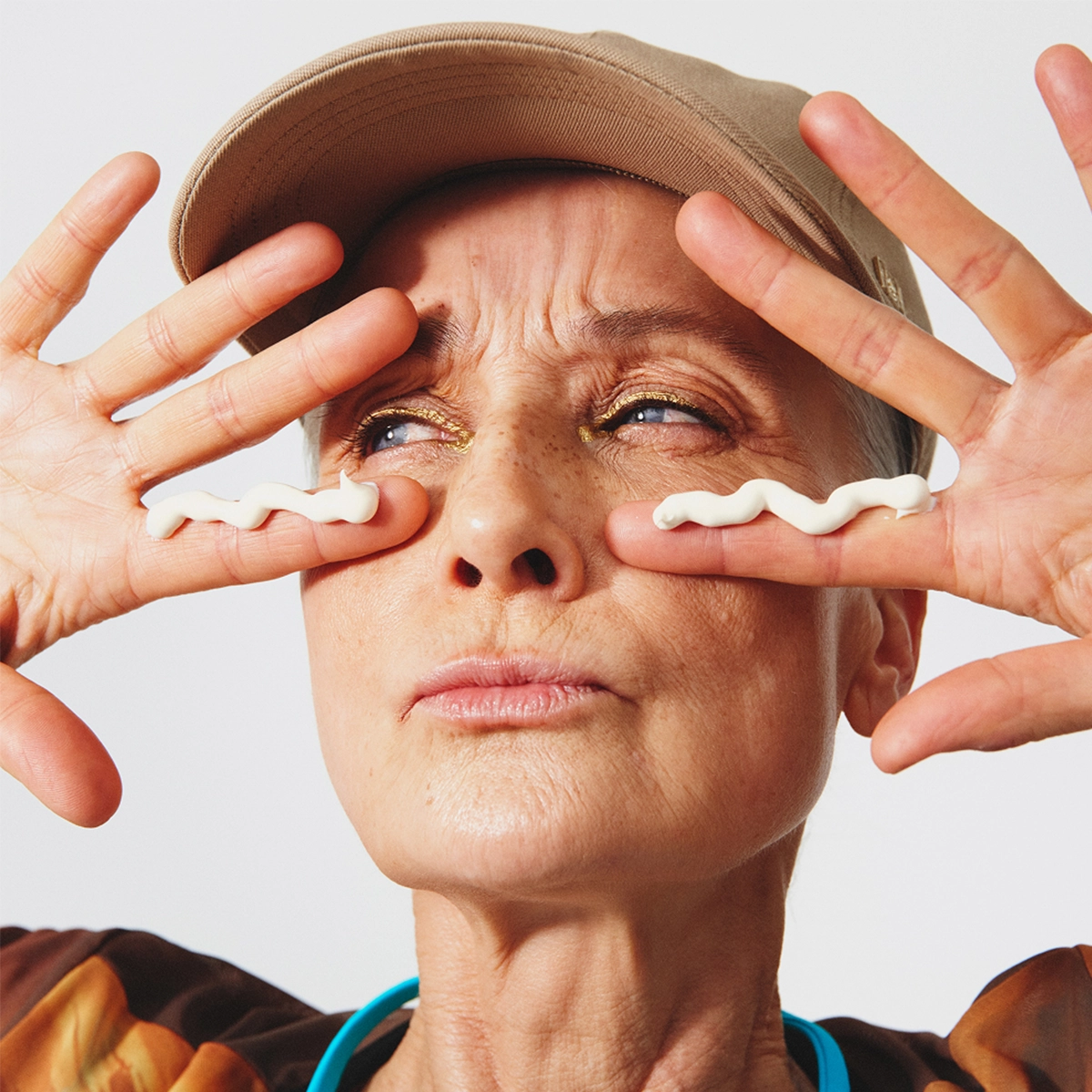 Muotokuva henkilöstä vaaleaa taustaa vasten. Henkilö pitää molempia käsiä kasvojensa edessä, ja vaaleaa voidetta on levitetty aaltoilevana viivana molempien silmien alle. Henkilöllä on lippalakki ja kullan sävyinen silmämeikki. Sormet ovat ojennettuina kasvojen molemmilla puolilla.