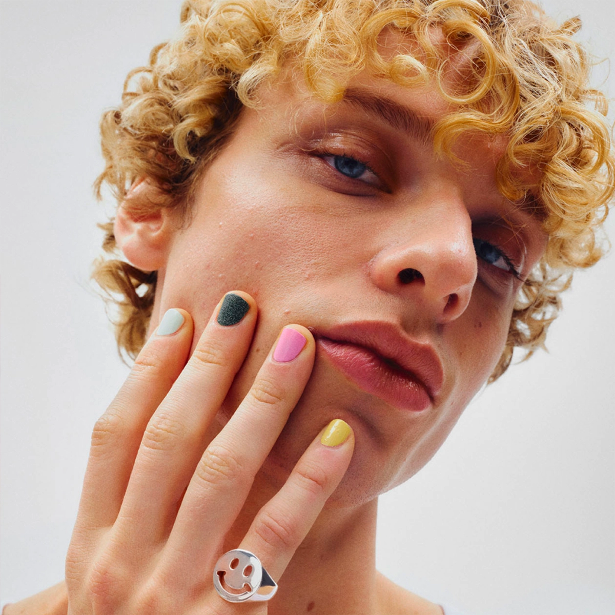 Nærbilde av ansikt med hånden mot kinnet og negler lakkert i ulike farger som rosa, gul, grønn og lyseblå. Kort krøllete hår, naturlig hudtekstur og nøytral bakgrunn.