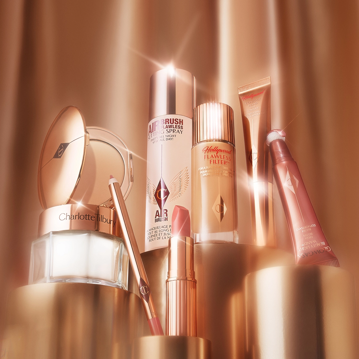 En samling av Charlotte Tilbury skjønnhetsprodukter, inkludert Airbrush Flawless Setting Spray, Hollywood Flawless Filter, Hollywood Contour Wand, Glowgasm Beauty Light Wand, en leppeblyant og en pudderkompakt. Produktene er elegant plassert og belyst med et varmt lys.