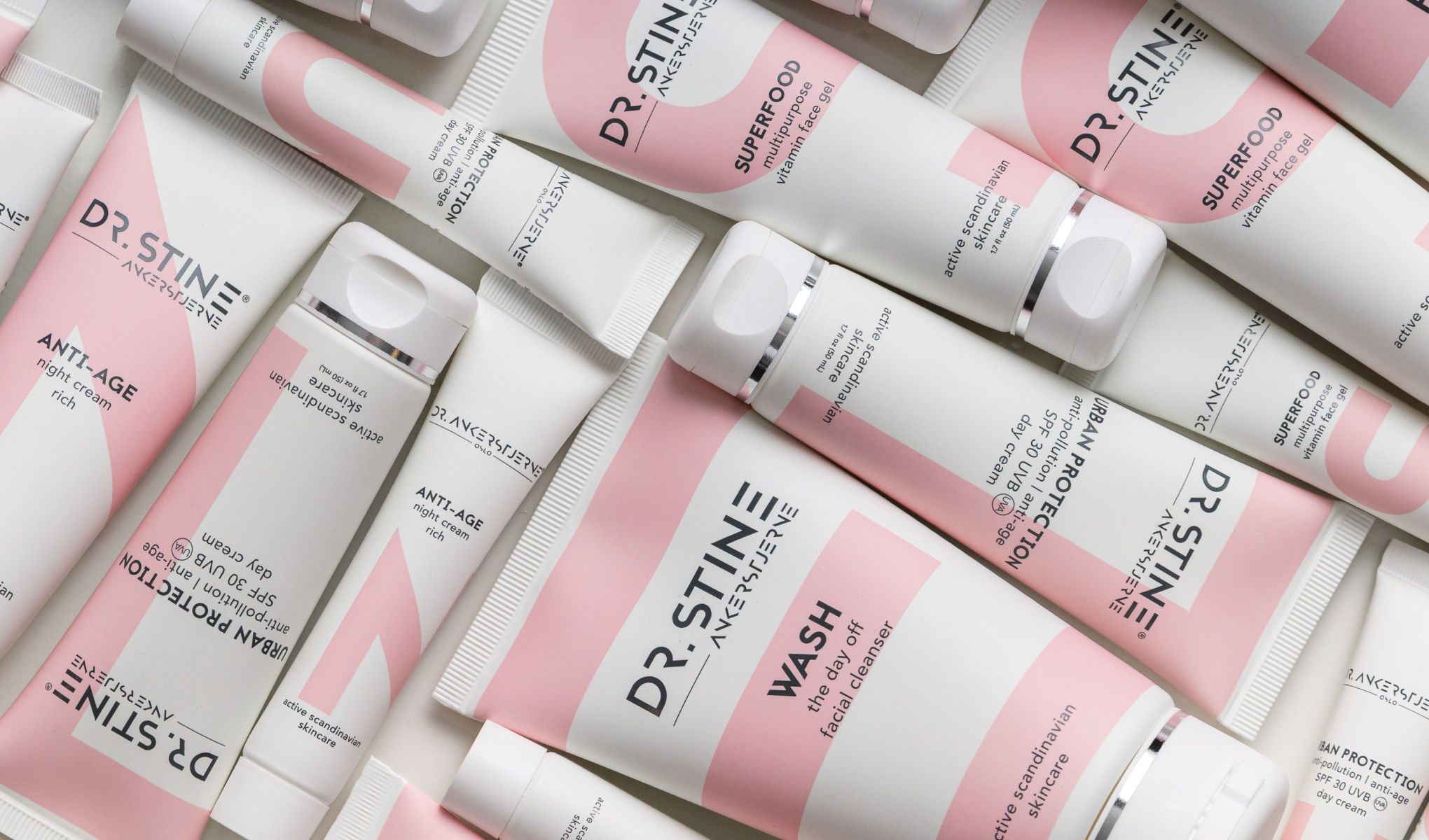 DR. STINE hudpleieprodukter i hvite og rosa emballasjer arrangert mot lys bakgrunn. Synlig tekst inkluderer “DR. STINE”, “SUPERFOOD multipurpose vitamin face gel”, “ANTI-AGE night cream rich”, “URBAN PROTECTION anti-pollution anti-age SPF 30 UVB/UVA day cream” og “WASH the day off facial cleanser”.