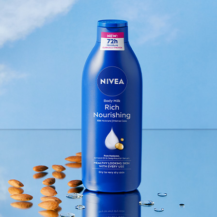 En blå flaske fra Nivea plassert mot en lyseblå bakgrunn. Flasken har teksten «NEW: 72h Moisture» og «Rich Nourishing» på etiketten. Rundt flasken er det mandler og noen vanndråper.