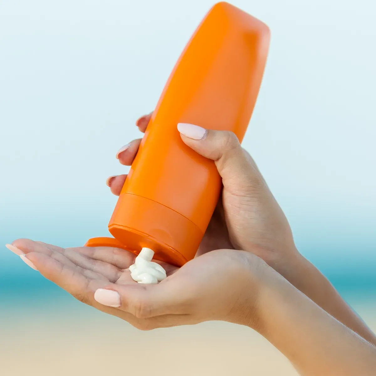 En person håller en orange flaska och applicerar solkräm på sin hand vid stranden.