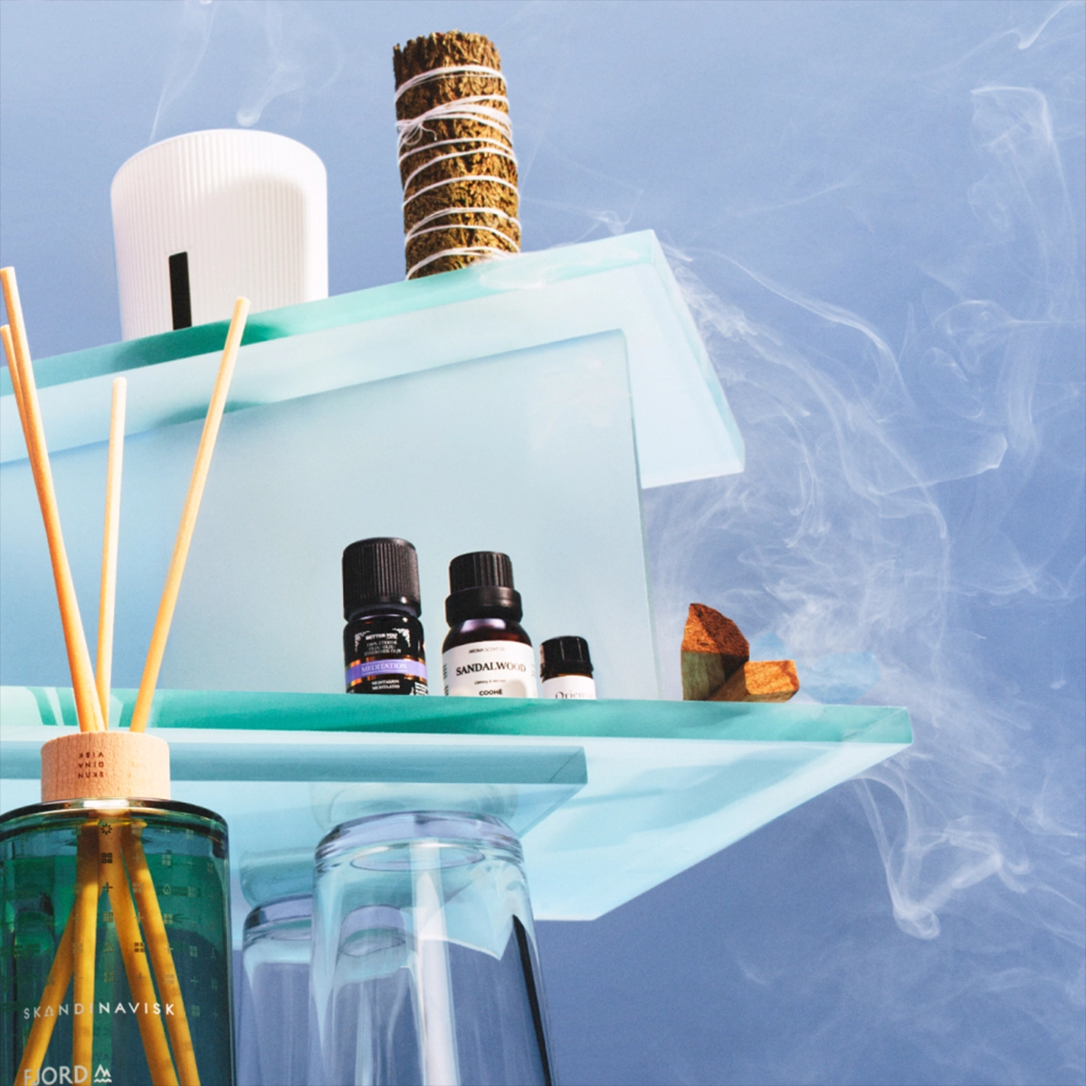 Doftprodukter och aromaterapi arrangerade på transparenta glasplattformar mot blå bakgrund med rökdetaljer. En diffuser från SKANDINAVISK FJORD med doftpinnar står bredvid eteriska oljor, bland annat “Sandalwood”, en vit diffuser och en bunt salvia för hemdoft och wellness.