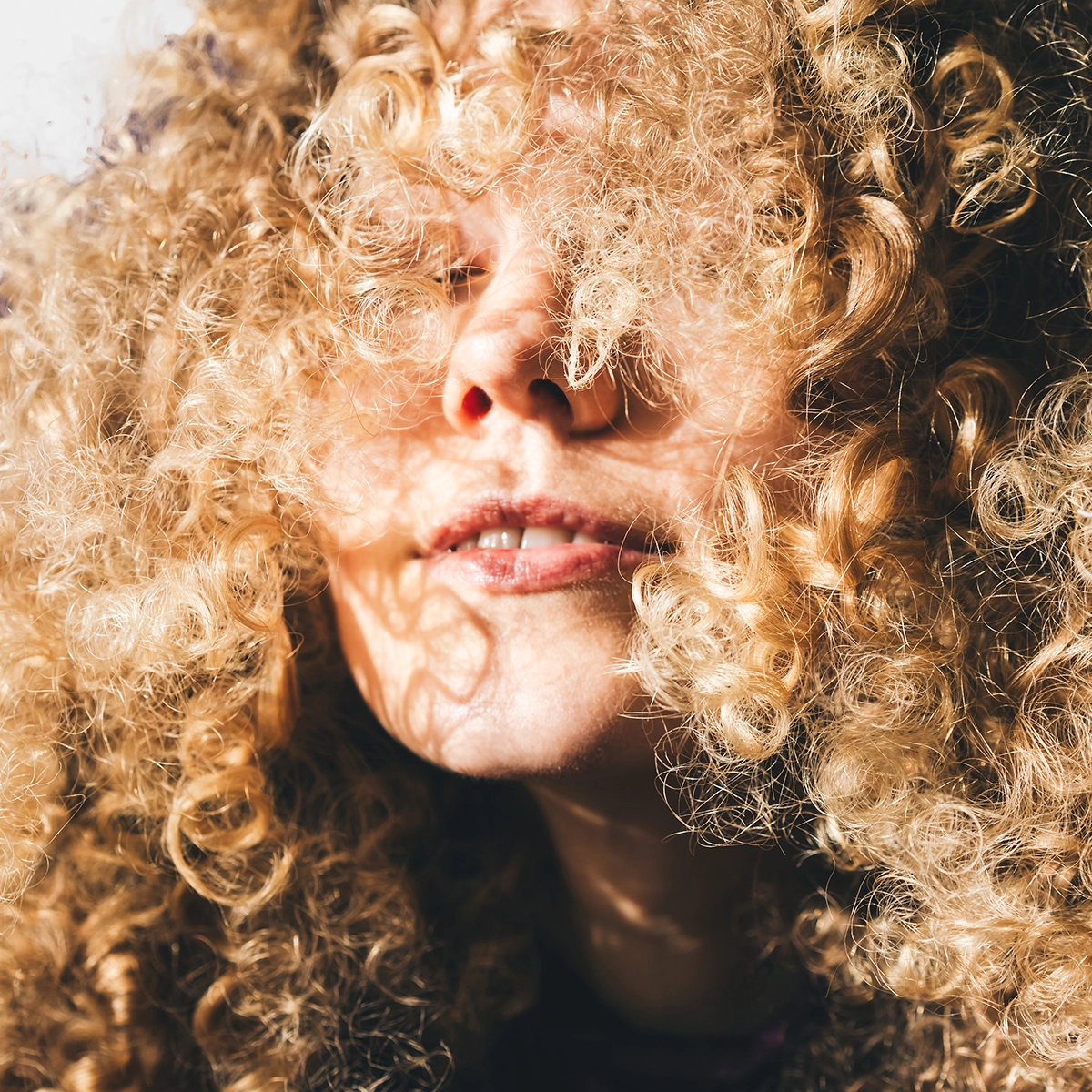 Nærbilde av en person med tette blonde krøller som dekker deler av ansiktet i varmt lys. Bildet fremhever naturlige krøller og hårtekstur for hårpleie og styling av krøllete hår.