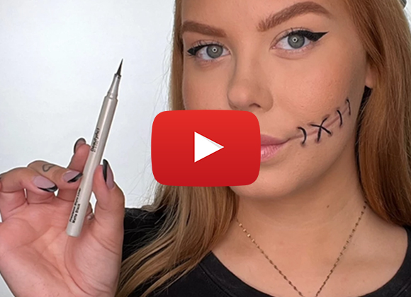 Person med blondt hår og falske sting på kinnet holder en eyelinerblyant. Personen har en tatovering på hånden og er iført en svart genser. Med et youtube play-symbol i midten av bildet 
