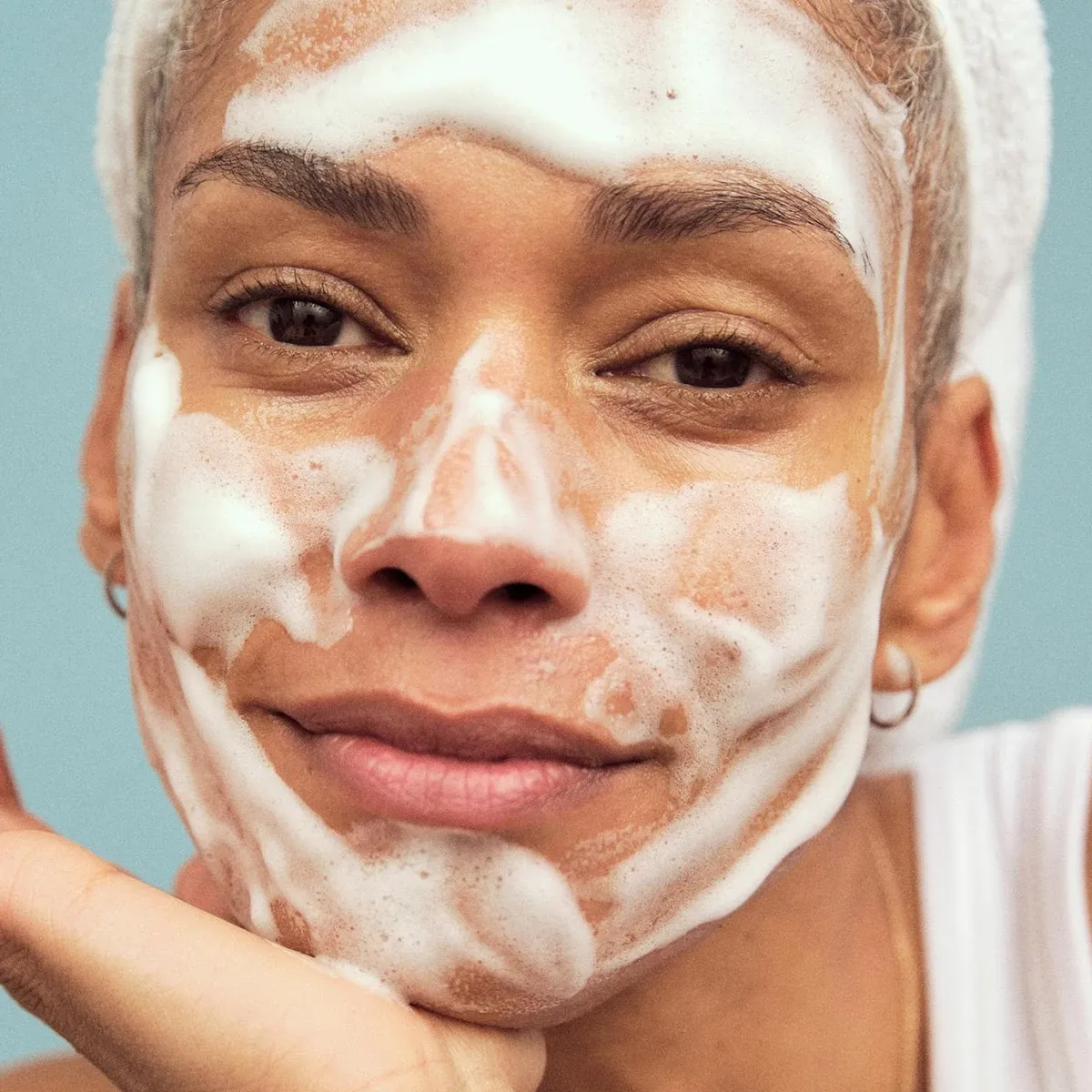 Närbild på kvinna med rengöringsskum i ansiktet, vilar hakan i handen och bär en vit handduk runt håret.