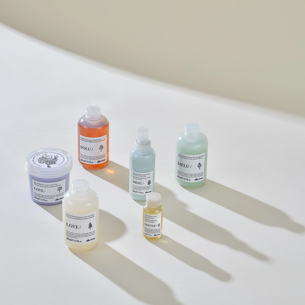 Davines hårvårdsprodukter från serierna LOVE, SOLU, MINU, MELU och NOUNOU står uppställda på vit yta med långa skuggor i mjukt ljus. Bilden visar schampo, hårmask, hårolja och värmeskyddsspray med produktnamn och Davines-logotyp synliga på förpackningarna.