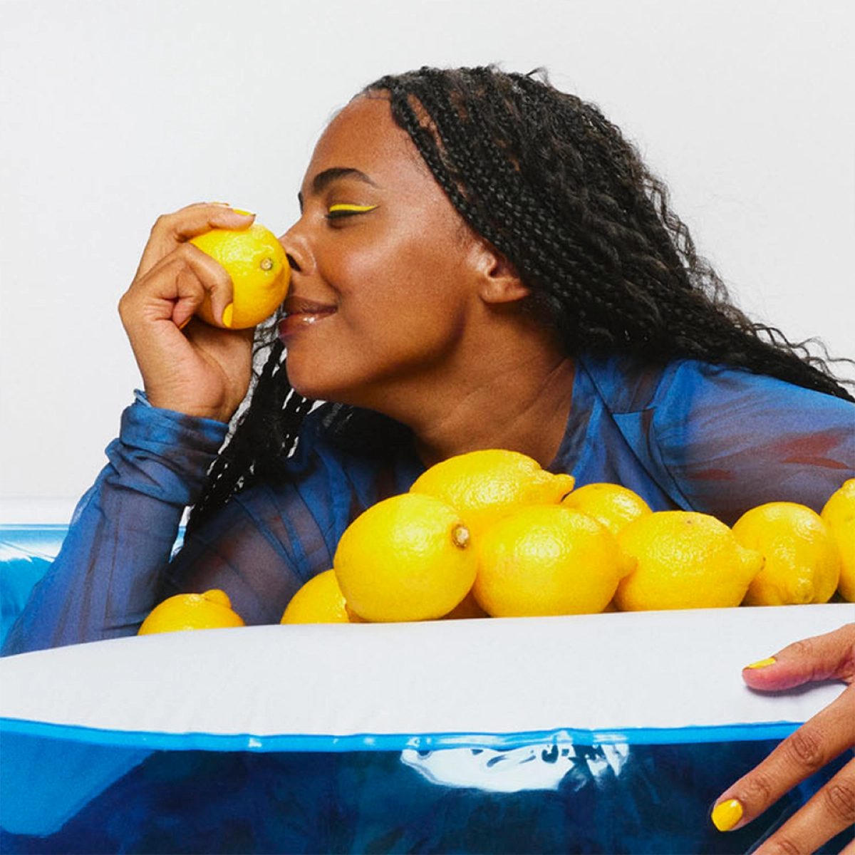 En person med långt lockigt hår håller en citron mot näsan. Personen bär en genomskinlig blå långärmad topp och lutar sig mot ett vitt kar fyllt med flera gula citroner. Bakgrunden är ljus och enfärgad.