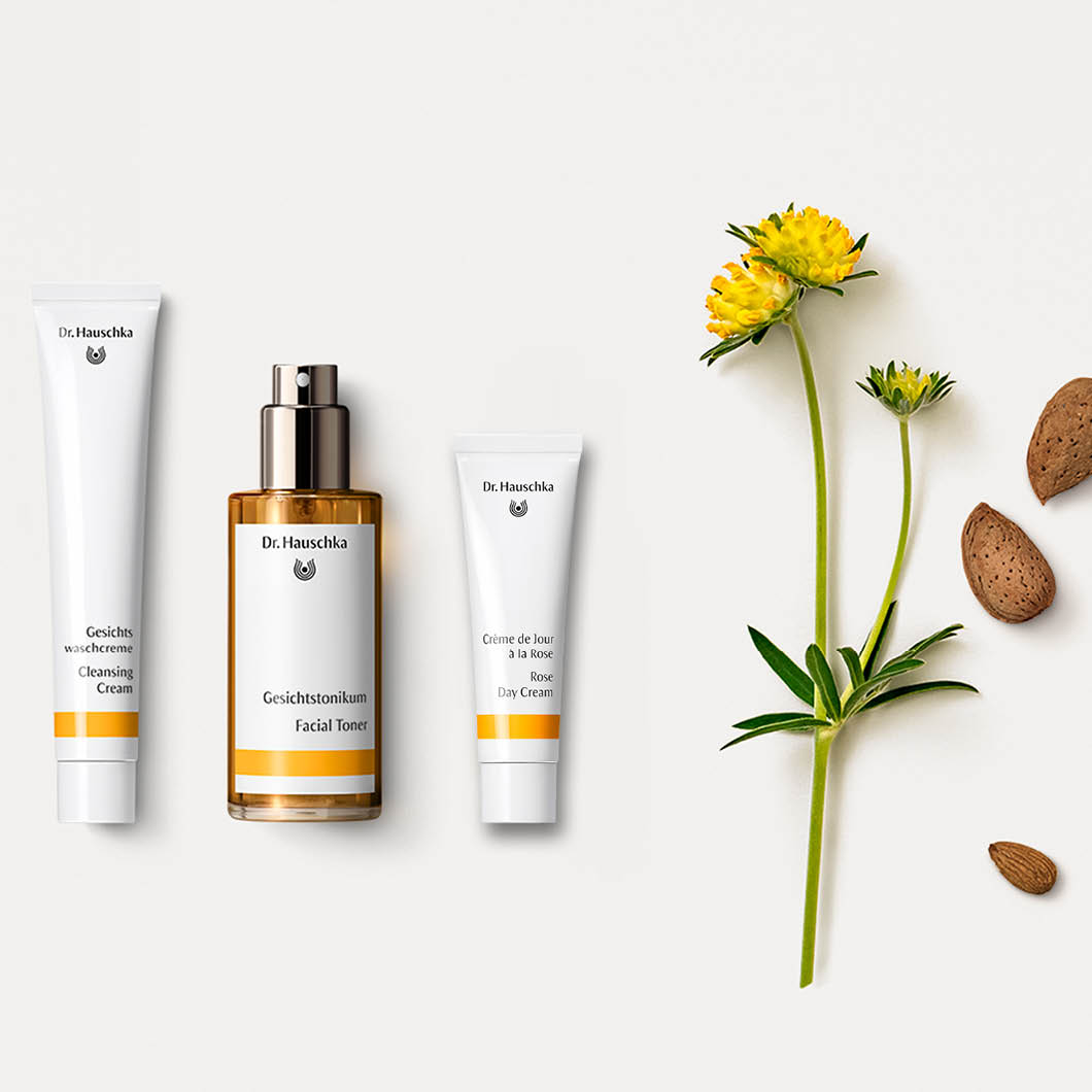 Tre Dr. Hauschka ansiktsprodukter med gul blomma och mandlar på vit bakgrund, ekologisk hudvård