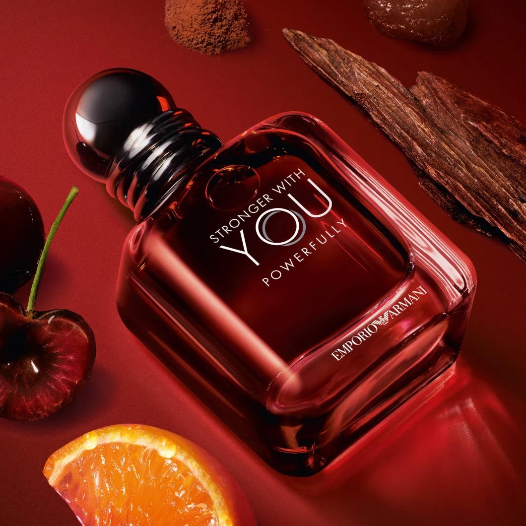 Emporio Armani Stronger With You Powerfully parfymflaska i mörkrött glas med texten "STRONGER WITH YOU POWERFULLY" och "EMPORIO ARMANI". Flaskan ligger på en röd bakgrund omgiven av körsbär, en apelsinklyfta, träbark och kryddor som förstärker doftens varma karaktär.