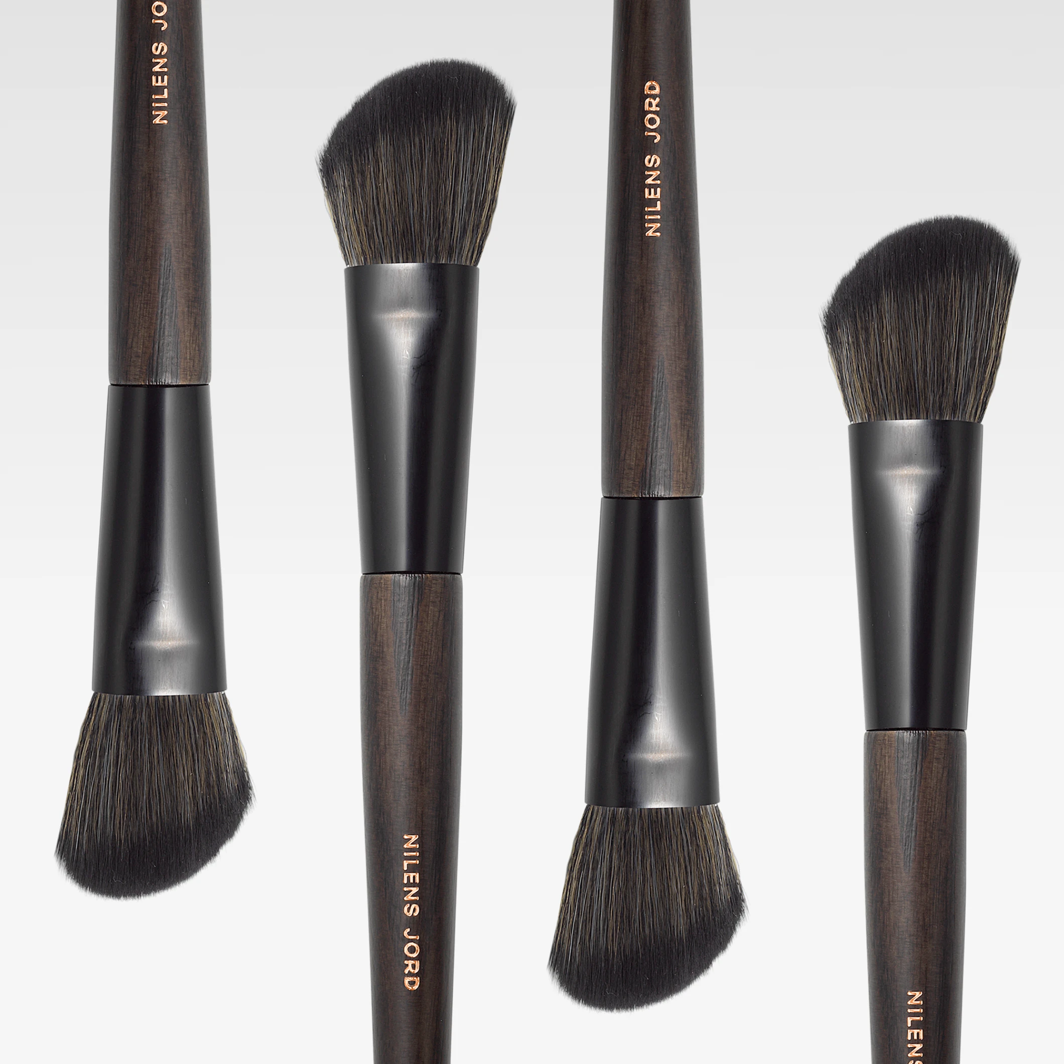Fyra sminkborstar från Nilens Jord med mörka trähandtag och svarta borsthuvuden, placerade vertikalt mot ljus bakgrund. Varumärkesnamnet ”Nilens Jord” syns på handtagen.