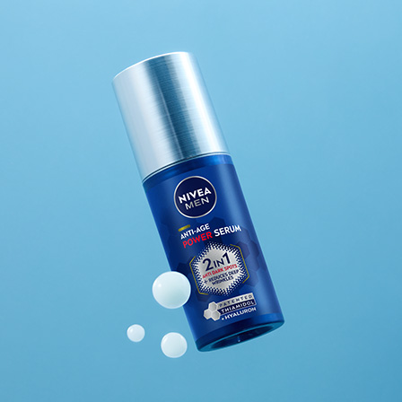 En blå flaske fra Nivea mot en lyseblå bakgrunn. Flasken har en sølvfarget pumpe og teksten «2in1 Intensive Serum» på etiketten. Rundt flasken er det noen hvite dråper av serumet, noe som understreker produktets tekstur.