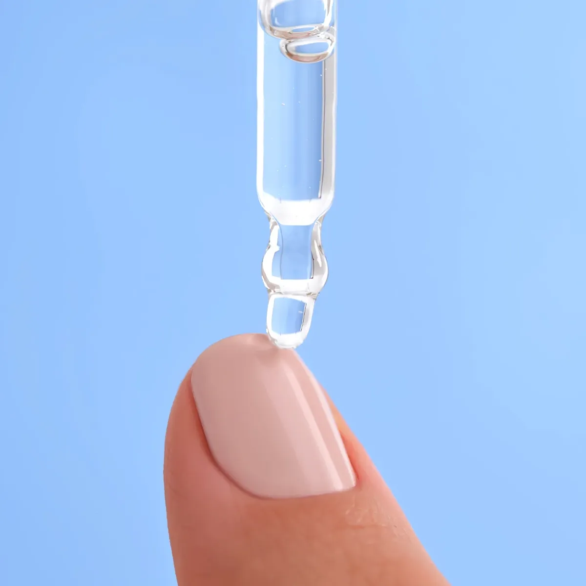 Nærbilde av en dråpe gjennomsiktig serum som påføres en velmanikyrert fingertupp mot en lys blå bakgrunn. Serumet dryppes fra en pipette, og fingertuppen har en lys rosa neglelakk.