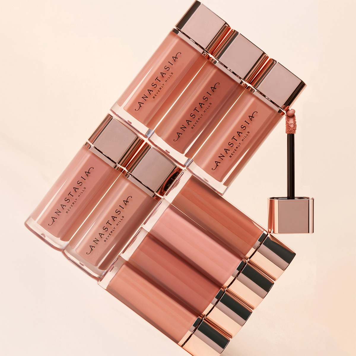 Bild på åtta läppglans från Anastasia Beverly Hills i olika nyanser av rosa och nude, arrangerade i ett geometriskt mönster. En av läppglansens applikatorer är utdragen och synlig.