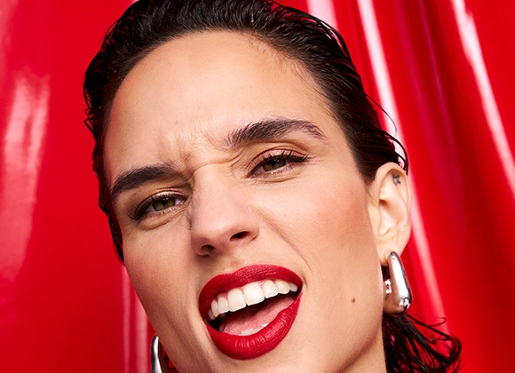 Närbild på en person med rött läppstift, bakåtkammat hår och stora silverörhängen mot en glansig röd bakgrund. En kraftfull och självsäker look som förmedlar festlig glamour och energi.