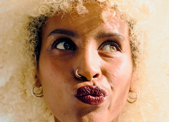 Närbild av en person med lockigt hår som tittar uppåt i sidled. Personen har markerade ögonbryn, mascara, mörkt läppstift, näsring och örhängen mot en ljus bakgrund.
