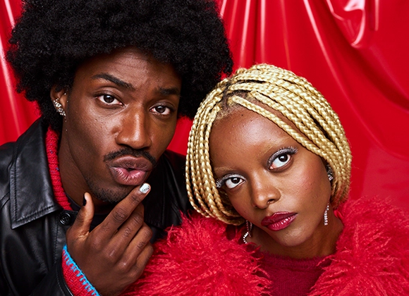 Bild som visar två personer med olika frisyrer och kläder. Den ena personen har mörkt lockigt hår och bär en svart läderjacka med en röd tröja. Den andra personen har blont flätat hår och bär en röd fuskpälsjacka. Bakgrunden är röd.