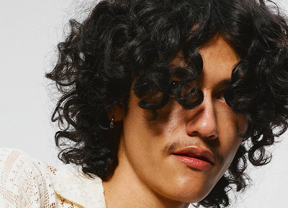 Lähikuva henkilöstä, jolla on tummat, kiharat hiukset otsalla ja kasvojen ympärillä. Henkilöllä on vaalea, teksturoitu paita ja taustana on neutraali pinta.