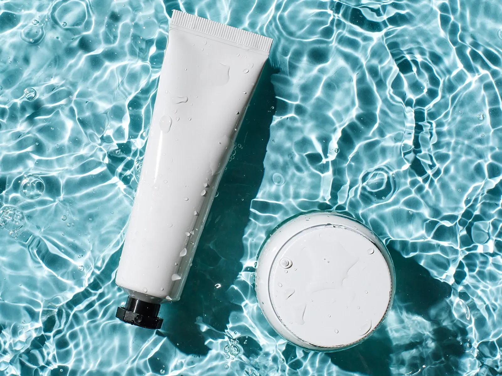 Hudvårdsprodukter – en tub och en burk – flytande i vatten med solljusreflektioner.