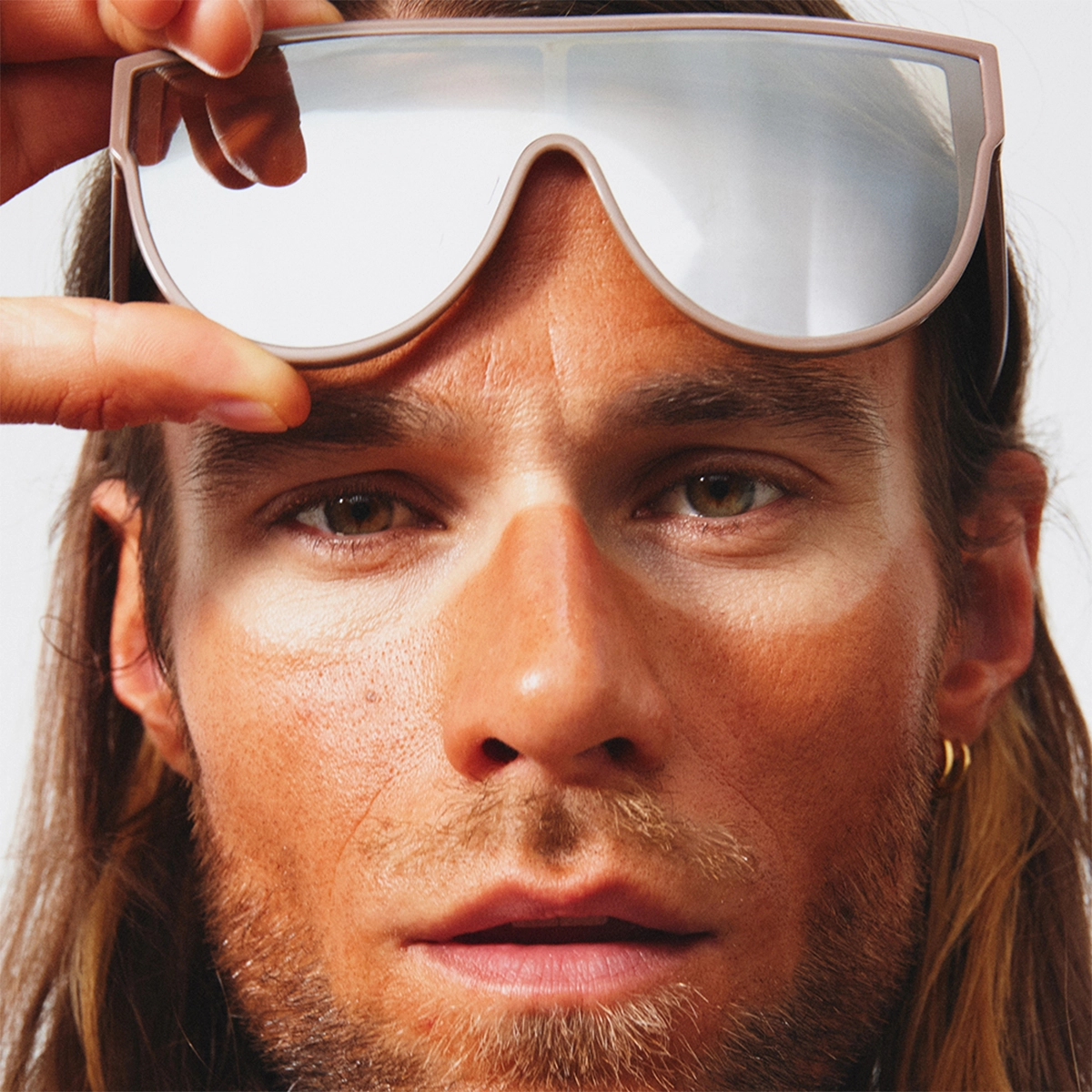 Lähikuva henkilöstä, joka nostaa ylös suuret peililinssiset aurinkolasit beigen värisillä kehyksillä. Kasvoilla on aurinkoinen, ruskea meikkipohja tai bronzer, ja silmien ympärillä näkyy selvästi vaaleampi iho aurinkolasien kohdalta.