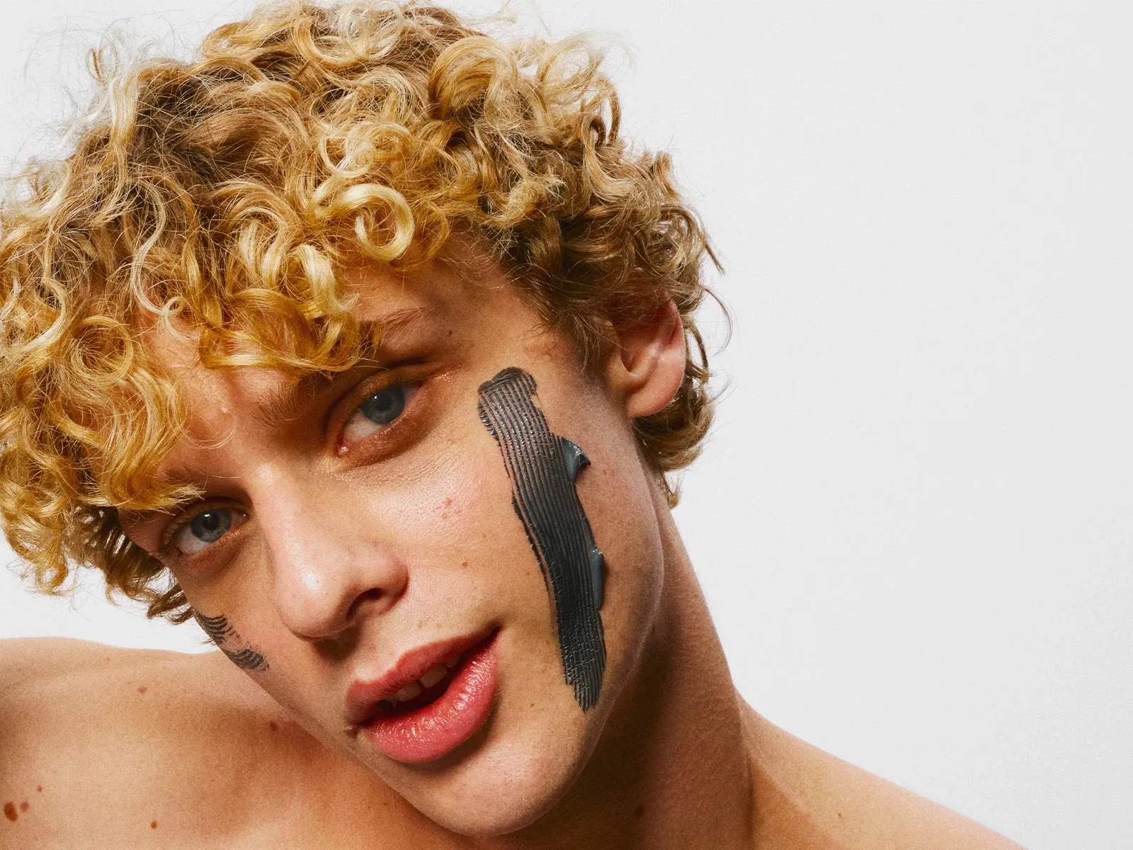 Henkilö, jolla on lyhyet, kiharat ja vaaleat hiukset sekä musta kasvonaamio levitettynä poskille, valkoista taustaa vasten.